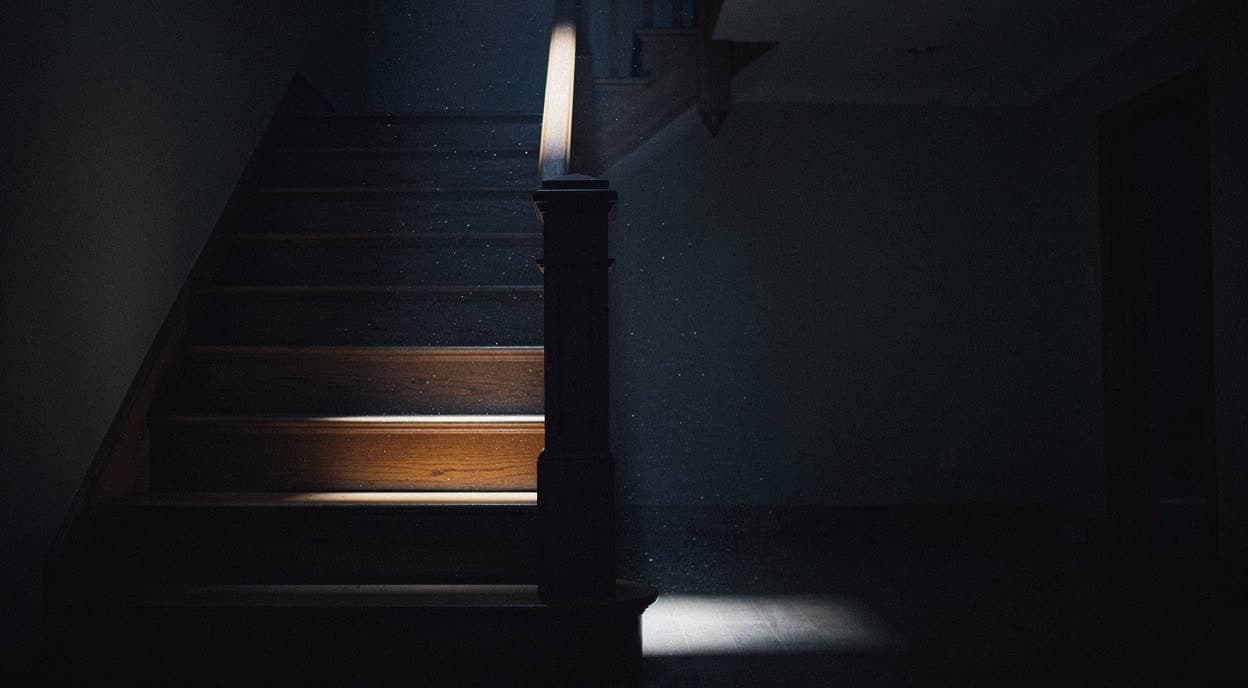 The foot of a wooden staircase in darkness, one tread catching a sliver of light